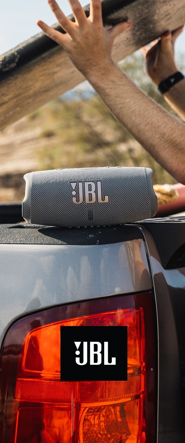 Enceinte portable JBL en tissu gris avec des accents en métal, positionnée sur le toit d'un véhicule, avec un logo sur le feu arrière en arrière-plan.
