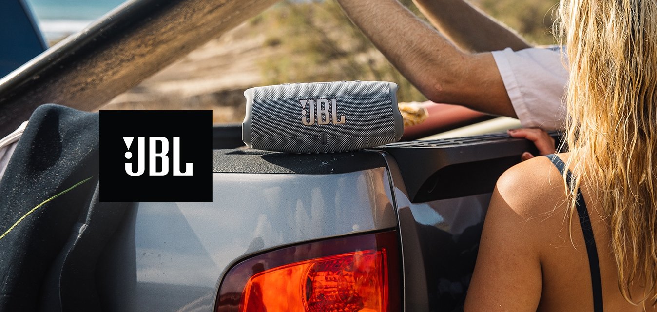 Šedý přenosný reproduktor JBL na ložné ploše auta, po stranách černý neopren a bílá košile s dlouhým rukávem. Surfovací prkno drženo nad hlavou, plážové prostředí.