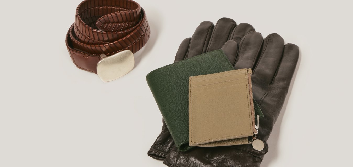 Skrutkový hnědý kožený pásek se stříbrnou sponou vedle černých kožených rukavic, na kterých je zelený pas a béžová peněženka, na bílé ploše.