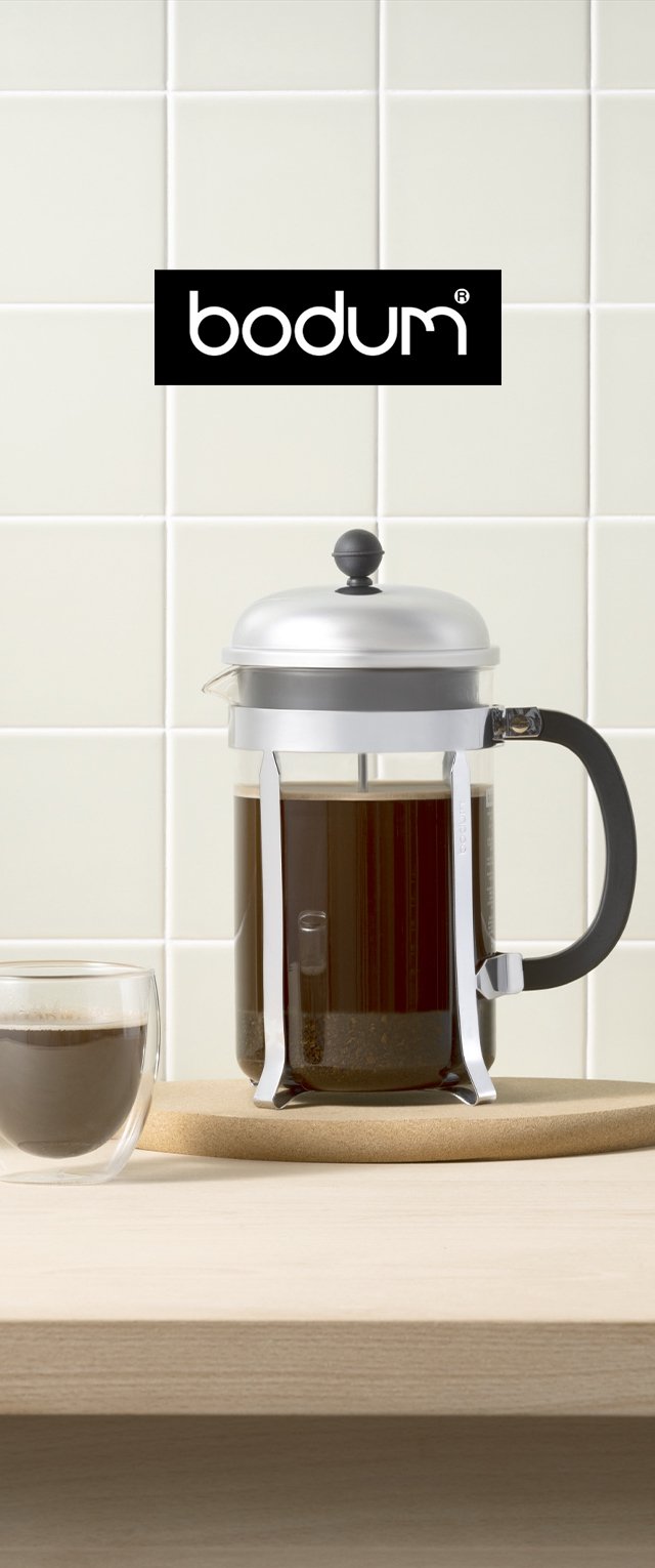 Prensa francesa Bodum con tapa de acero inoxidable y mango negro, sosteniendo café oscuro. Taza de vidrio transparente al lado sobre una superficie de madera.