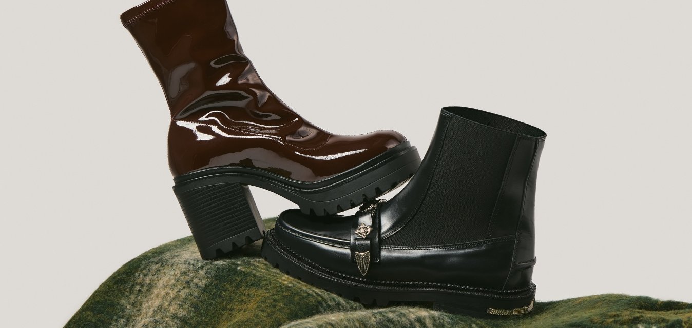 Hnědá kotníková obuv na podpatku z lakované kůže a černá kožená kotníková obuv s přezkou položené na složené zelené vzorované vlněné šále.