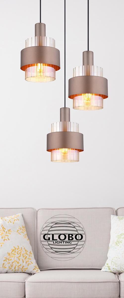 Trzy lampy wiszące z metalowymi kloszami i przezroczystymi cylindrycznymi akcentami, zawieszone na czarnych przewodach, nad jasnoszarym sofą z wzorzystymi poduszkami.