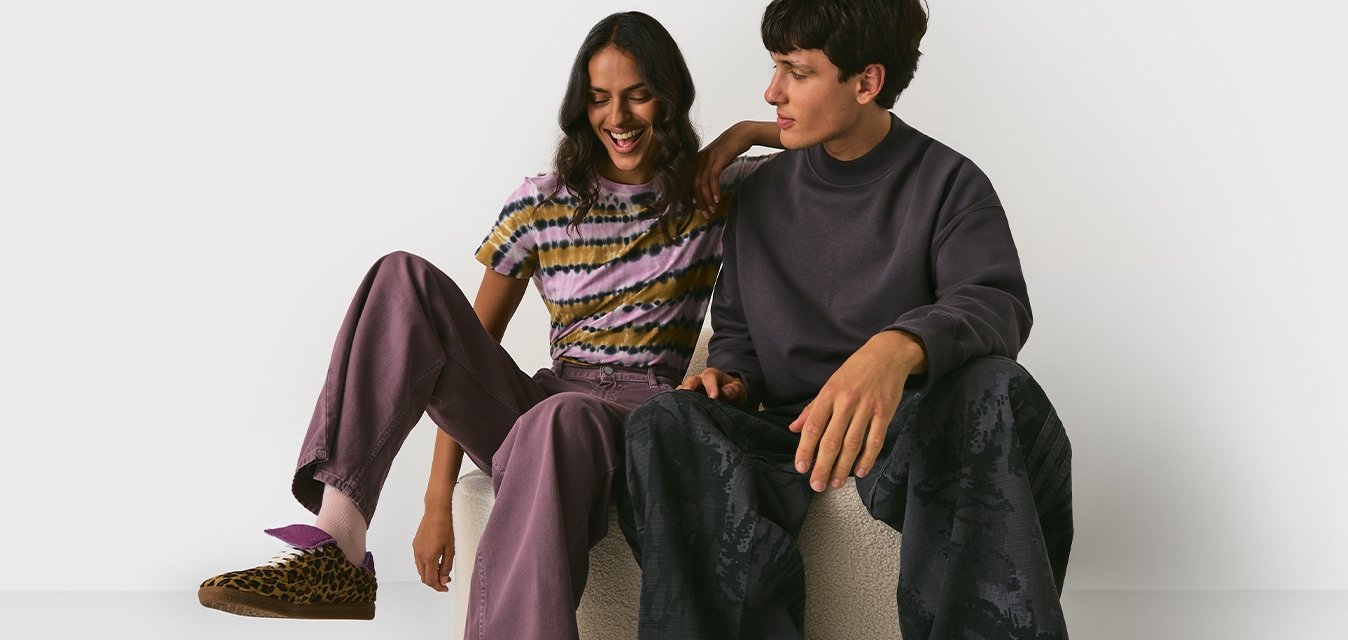 Dva mladí dospělí v neformálním oblečení sedící na světlém otomanu, usmívající se, na jednobarevném bílém pozadí.