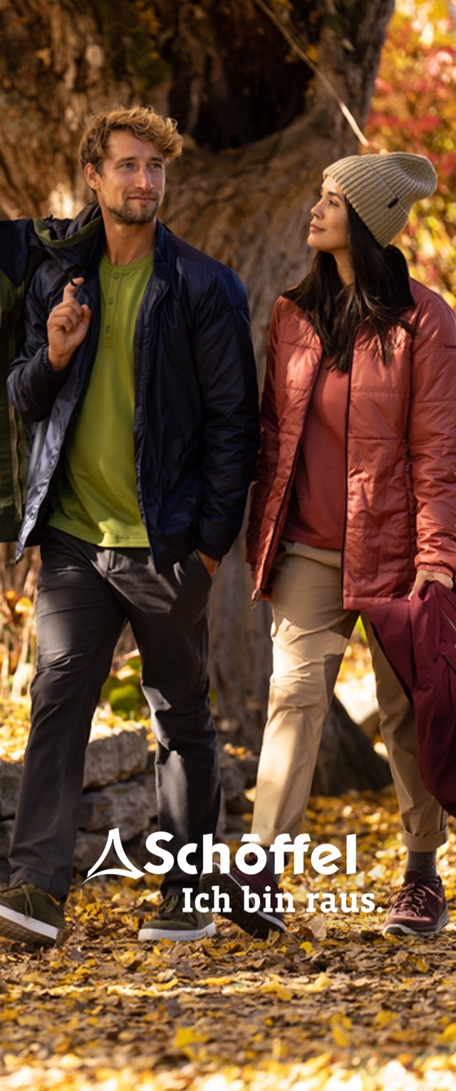 Dos personas caminan por un sendero rodeado de hojas de otoño. Uno lleva una chaqueta azul marino y una camisa verde, el otro una chaqueta burdeos y pantalones beige.