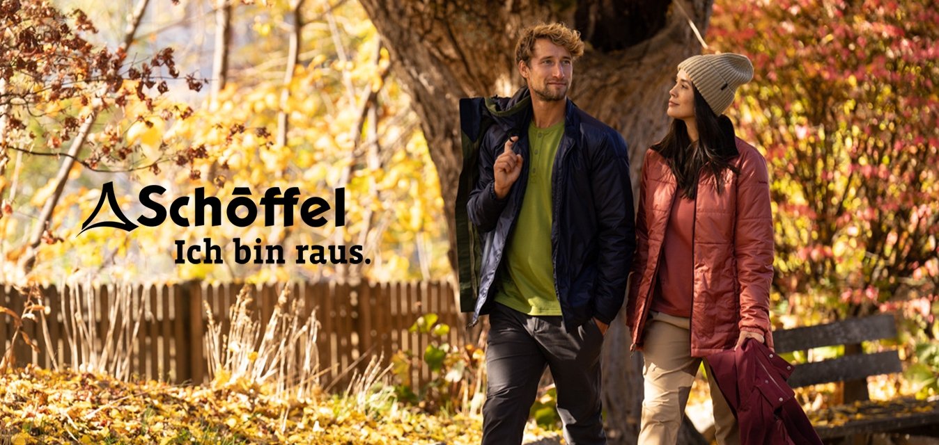 Dva lidé v bundičkách kráčejí po cestičce pokryté listím v parku se stromy v podzimních barvách a poblíž je dřevěný plot.