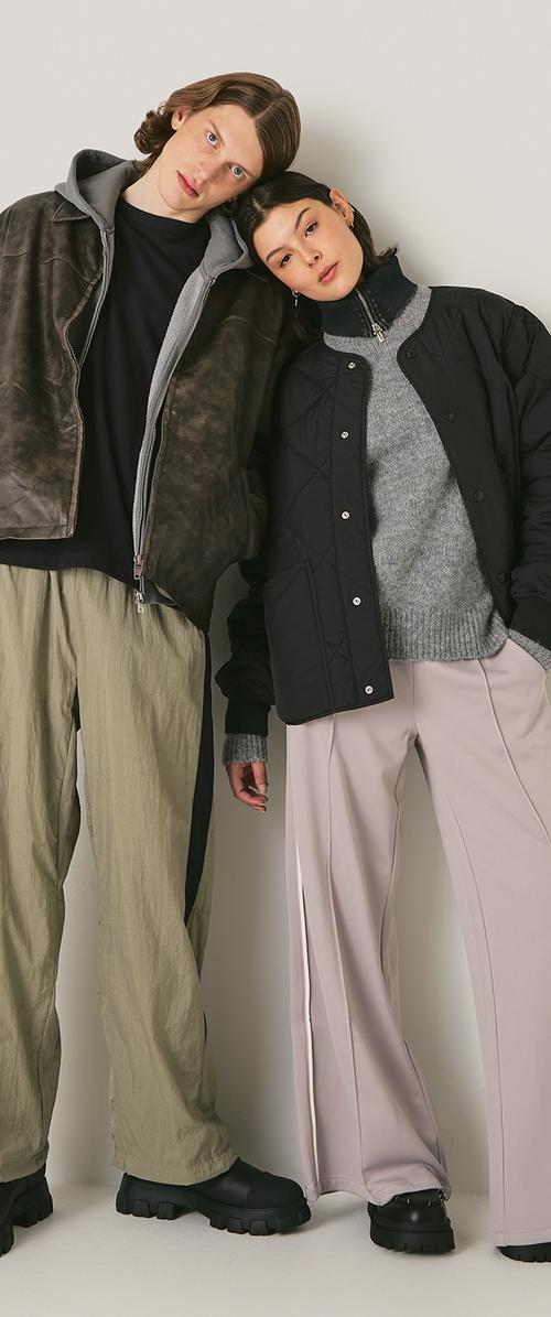 Dos modelos se apoyan uno en el otro. Uno lleva una chaqueta bomber oscura, un suéter gris claro y pantalones verde claro. El otro tiene una chaqueta acolchada negra y pantalones rosa claro. Ambos llevan calzado negro grueso.