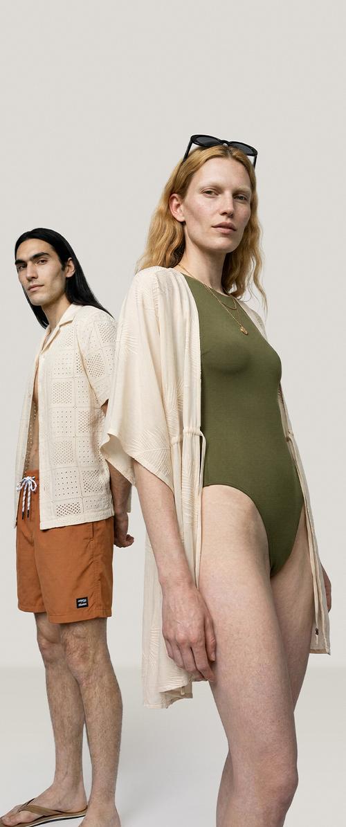 Zwei Models stehen Rücken an Rücken in Sommerkleidung; eines in einem olivfarbenen Badeanzug mit leichtem Überwurf, das andere in orangen Shorts und einem beigen offenen Hemd.