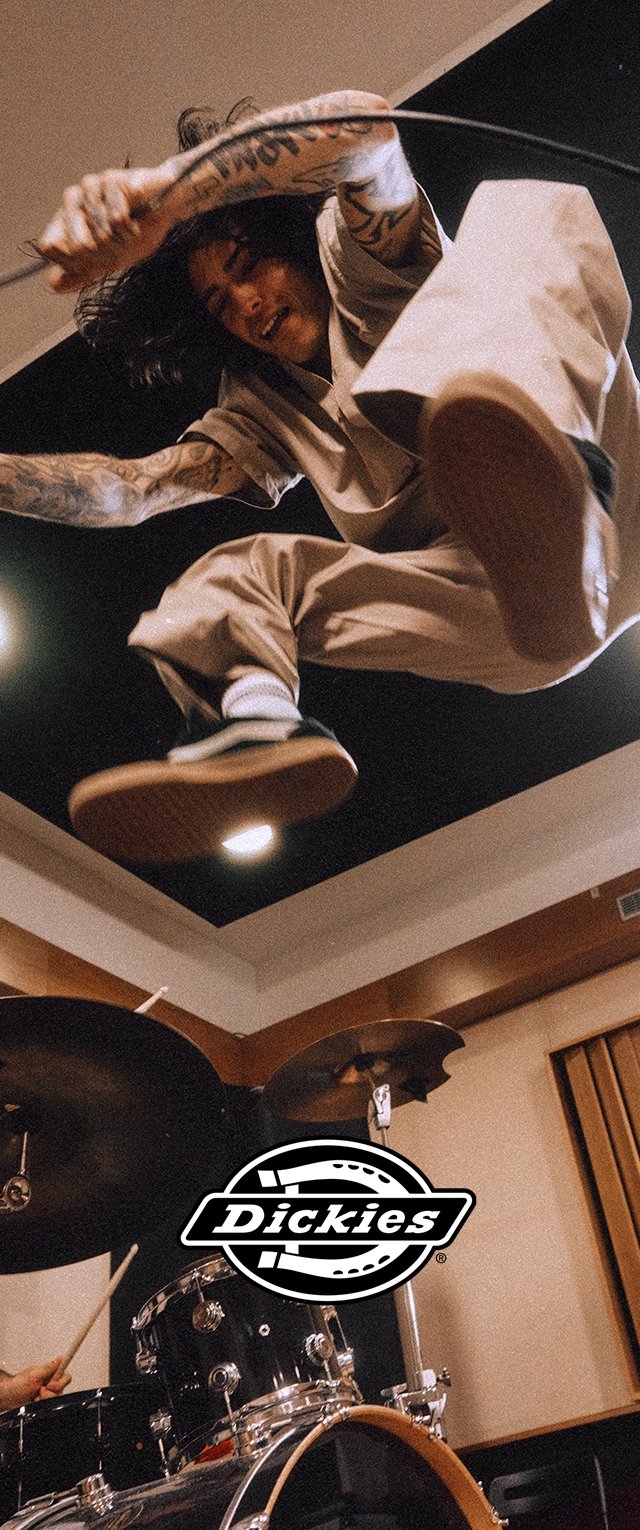 Una persona con un atuendo beige está saltando en el aire con una baqueta, frente a un fondo oscuro y paneles de madera, con una batería abajo.