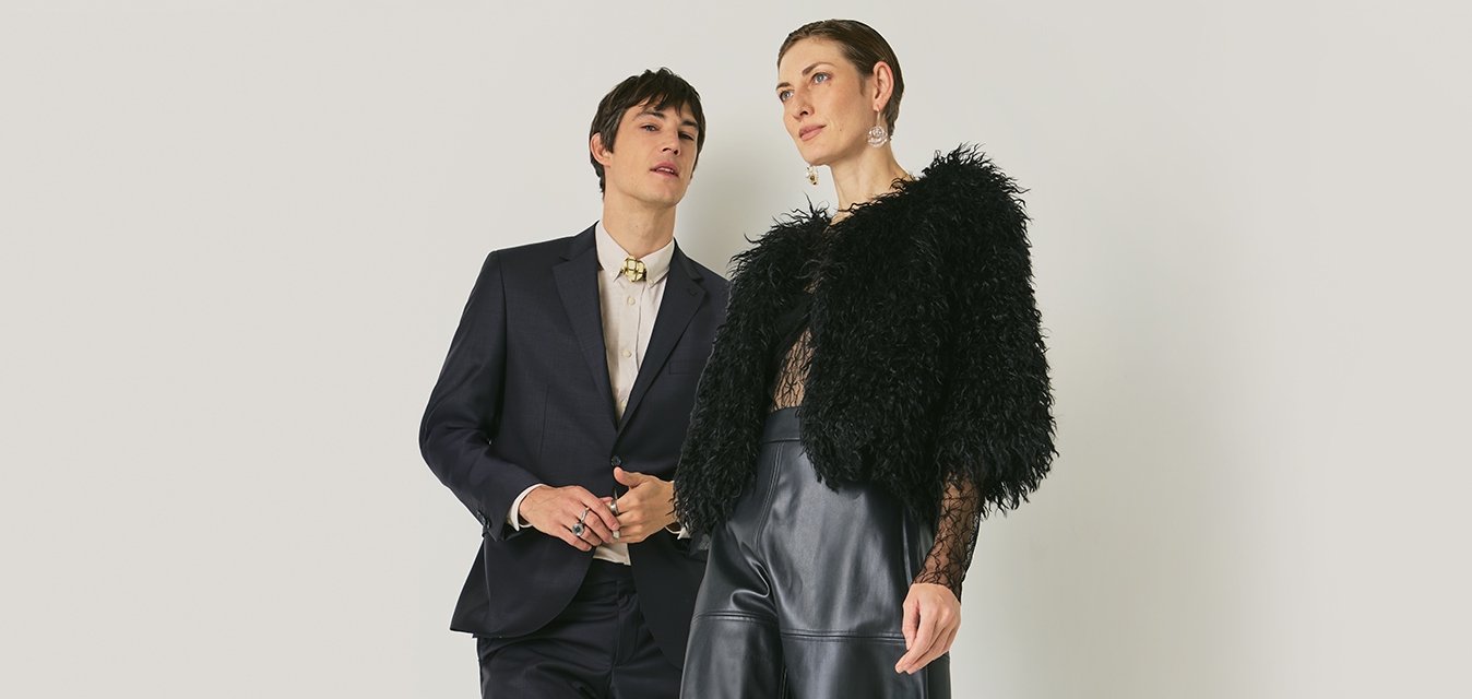 Dva modely stojí proti jednoduchému pozadí; jeden má na sobě tmavý oblek, druhý černou umělou kožich, krajkový top, koženou sukni a boty na podpatku.
