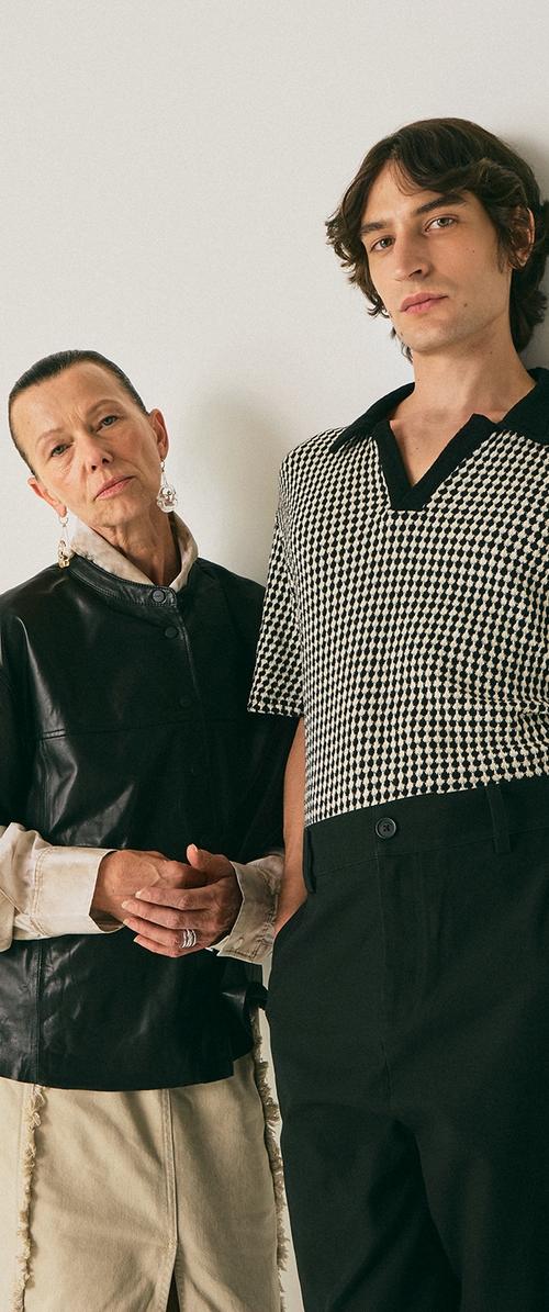 Une femme âgée en haut en cuir noir et jupe beige se tient à côté d'un jeune homme portant une chemise à motifs noir et blanc à manches courtes et un pantalon noir, sur un fond uni.