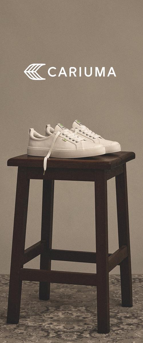 Para białych sneakersów Cariuma z rozwiązanymi sznurowadłami, umieszczona na ciemnym drewnianym stołku na jednolitym beżowym tle.
