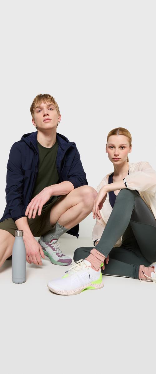 Twee mensen zitten op een lichte achtergrond. De man draagt een marineblauwe jas en groene shorts, terwijl de vrouw een beige jas en donkere leggings heeft. Een grijze waterfles en sportschoenen met groene accenten liggen in de buurt.