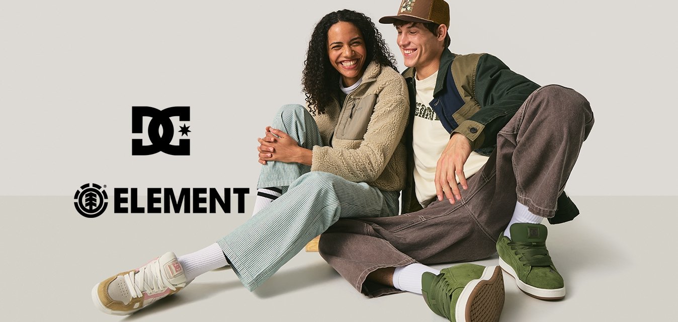 Dva lidé sedící na zemi, jeden má na sobě béžovou flísovou bundu, zelené manžestrové kalhoty a zelené tenisky, druhý hnědou čepici a modrou bundu.