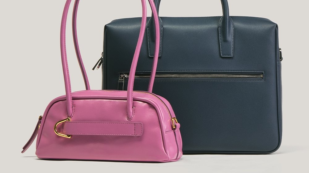 Zwei Lederhandtaschen: eine kleine rosa mit goldenen Beschlägen und langen Griffen, eine große dunkelblaue mit vorderer Reißverschlusstasche, beide aufrecht vor einfachem Hintergrund.