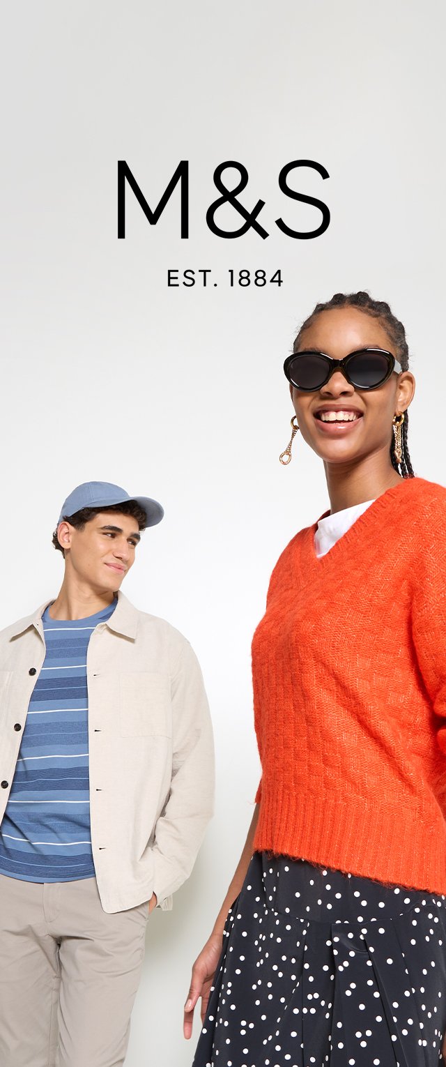 Pull en tricot orange, jupe noire à motifs avec des pois blancs, veste crème, chemise rayée bleue et casquette bleue. Modèle posé devant un fond blanc.