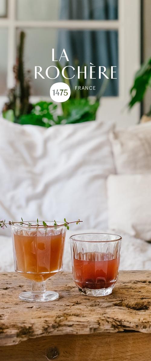Deux verres en verre sur une surface en bois ; l'un est haut avec un motif hexagonal, rempli d'un liquide orange ; l'autre est plus court, rond avec une boisson rougeâtre.