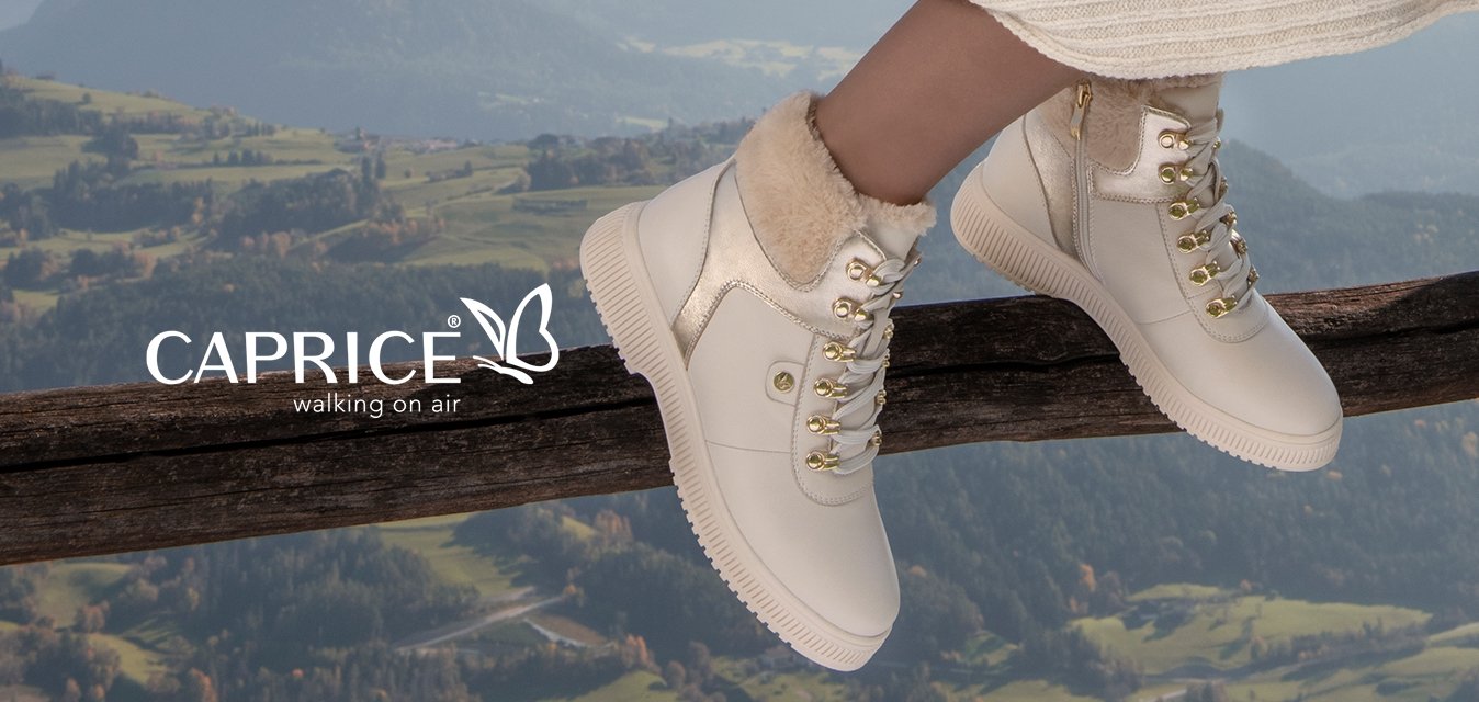 Pletený béžový cardigan, bílá žebrovaná šaty, bílé kotníkové boty s kožešinovou podšívkou a zlatými detaily, sedící na dřevěném zábradlí s horským pozadím.