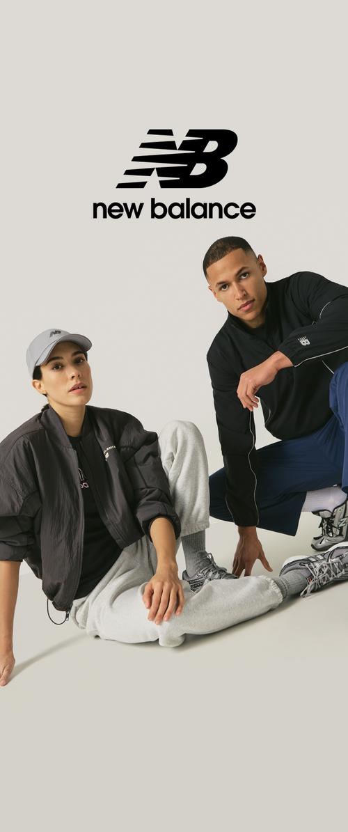 Chaqueta bomber negra, pantalones deportivos grises, camiseta deportiva negra y gorra. Las zapatillas presentan una combinación de gris, negro y rojo. Logo de New Balance visible.