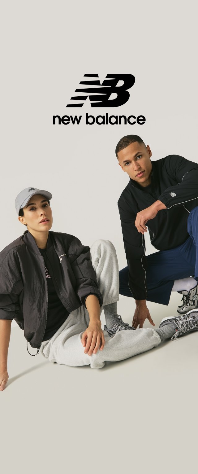 Chaqueta bomber negra, pantalones deportivos grises, camiseta deportiva negra y gorra. Las zapatillas presentan una combinación de gris, negro y rojo. Logo de New Balance visible.
