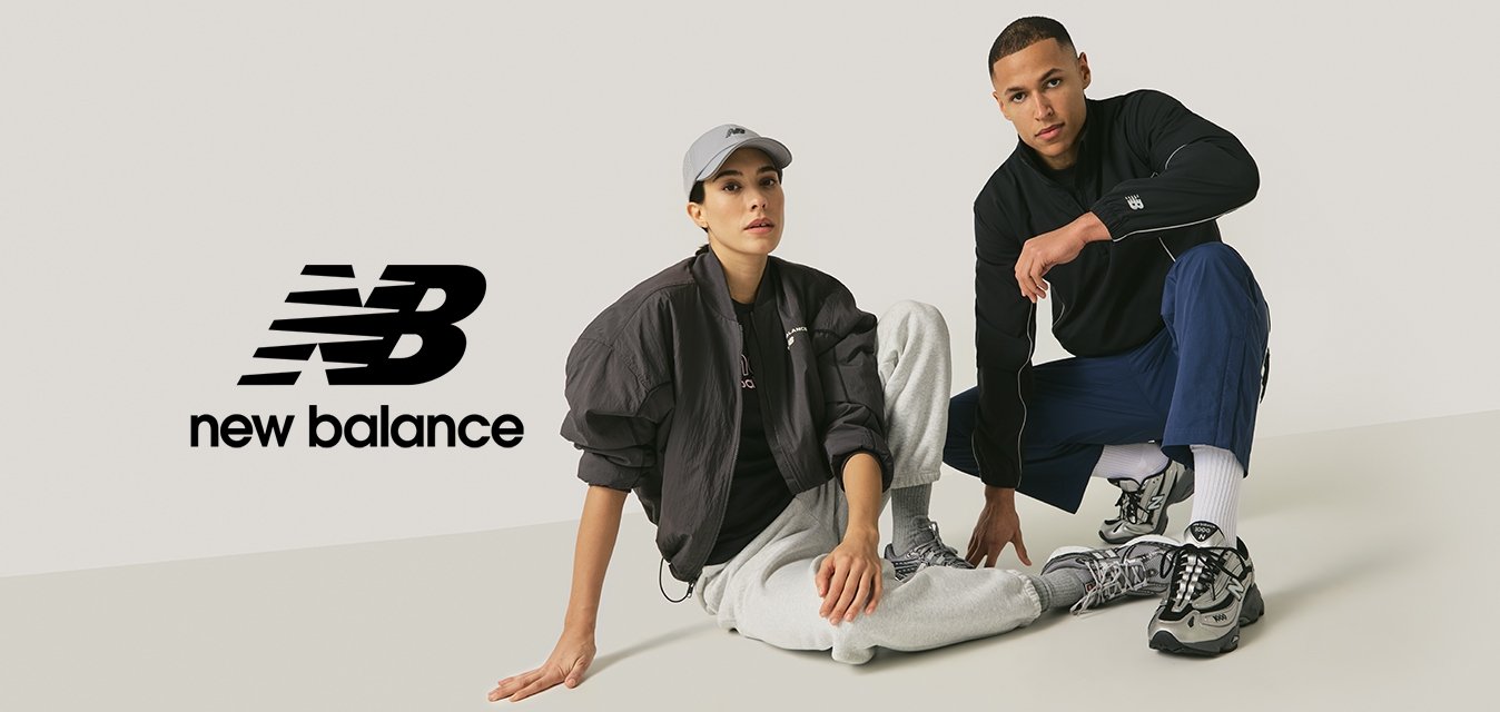 Žena v šedé čepici, černé bundě a šedých teplácích sedí vedle muže v černé bundě a modrých kalhotách, oba mají na nohou šedé tenisky na jednobarevném pozadí.