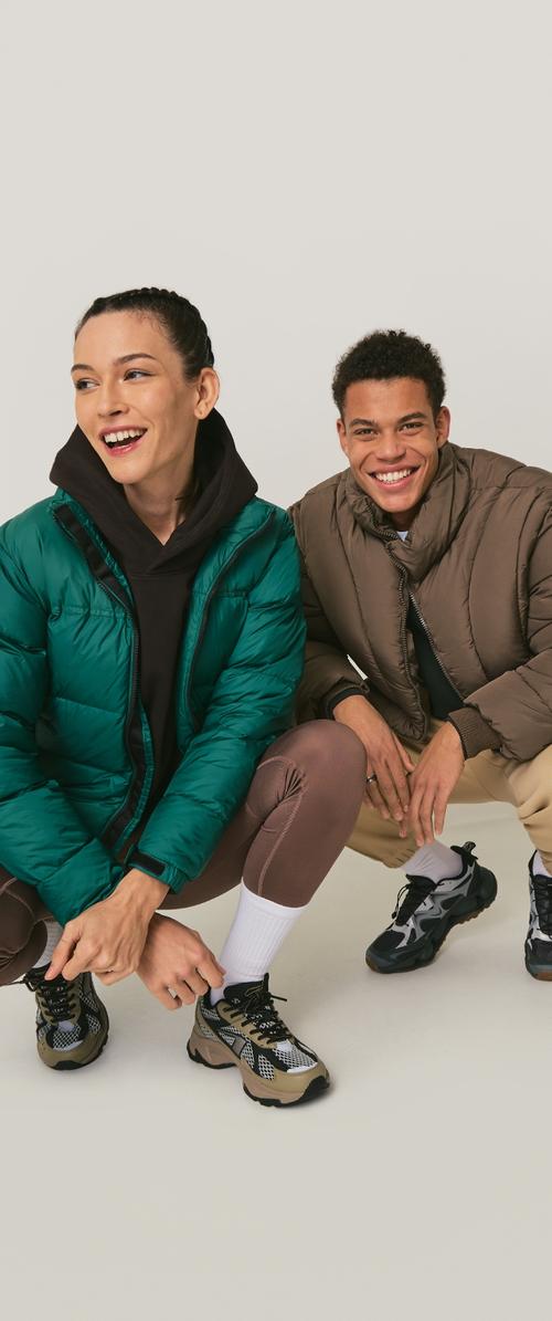 Doudoune verte portée sur un sweat à capuche noir, doudoune marron, leggings marron et baskets avec des détails en mesh ; tous deux accroupis devant un fond neutre.