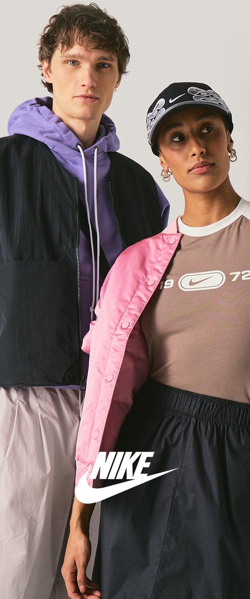 Jonge man in een paarse hoodie en zwart vest en jonge vrouw in een roze jas, beige Nike-shirt, zwarte rok en zwarte pet staan naast elkaar poseren.