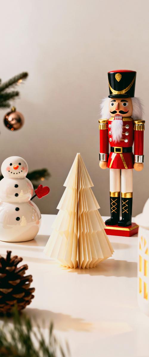 Schiaccianoci decorativo, albero di Natale di carta e pupazzo di neve in ceramica con guanto rosso su superficie bianca vicino a una pigna e ramo d'albero sfocato.