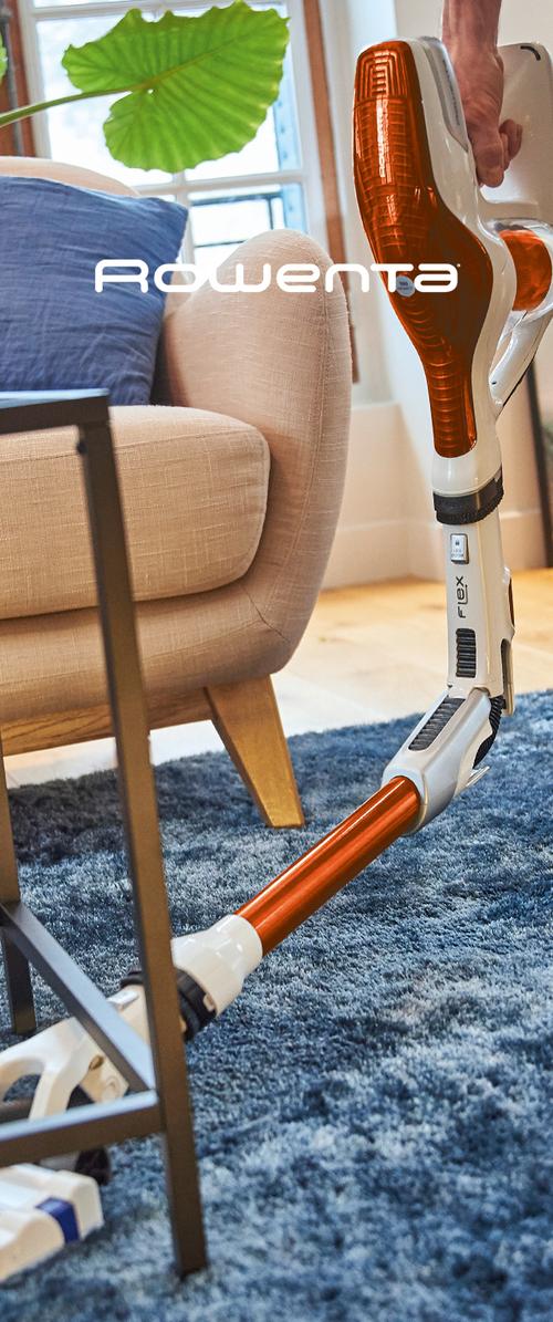 Draadloze stofzuiger met een wit en oranje ontwerp, met een flexibele scharnier, geplaatst naast een beige fauteuil op een blauwe vloerbedekking.