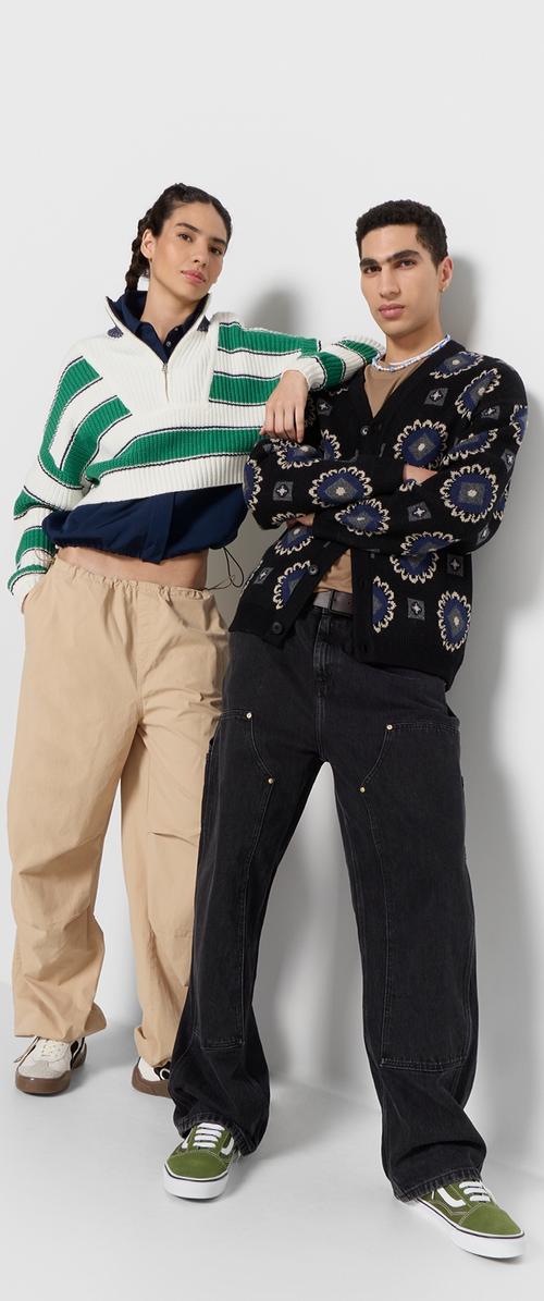 Brede gebreide trui met groene en witte strepen; zwarte cardigan met bloemmotieven; losse beige broek; zwart en groene sneakers. Casual look.