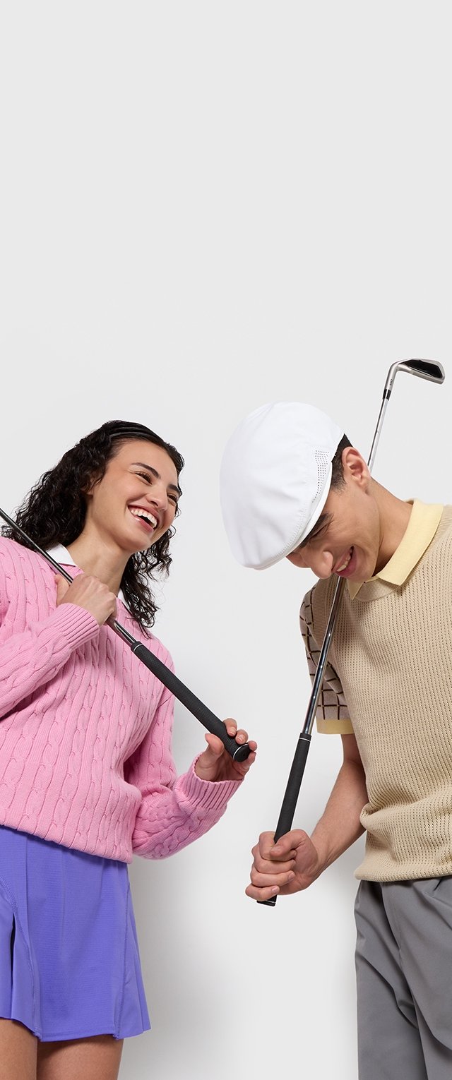 Dos jóvenes adultos sostienen palos de golf, vistiendo un suéter rosa de punto trenzado, una polo beige de punto y una gorra blanca, con un fondo neutral.