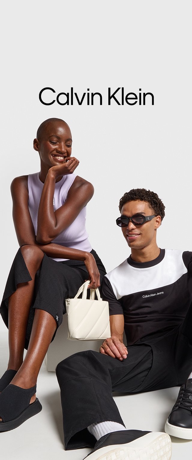 Dos personas están sentadas juntas. Una lleva una blusa de canalé lila. La otra tiene una camisa en blanco y negro, gafas de sol y pantalones negros. Hay un bolso blanco presente.