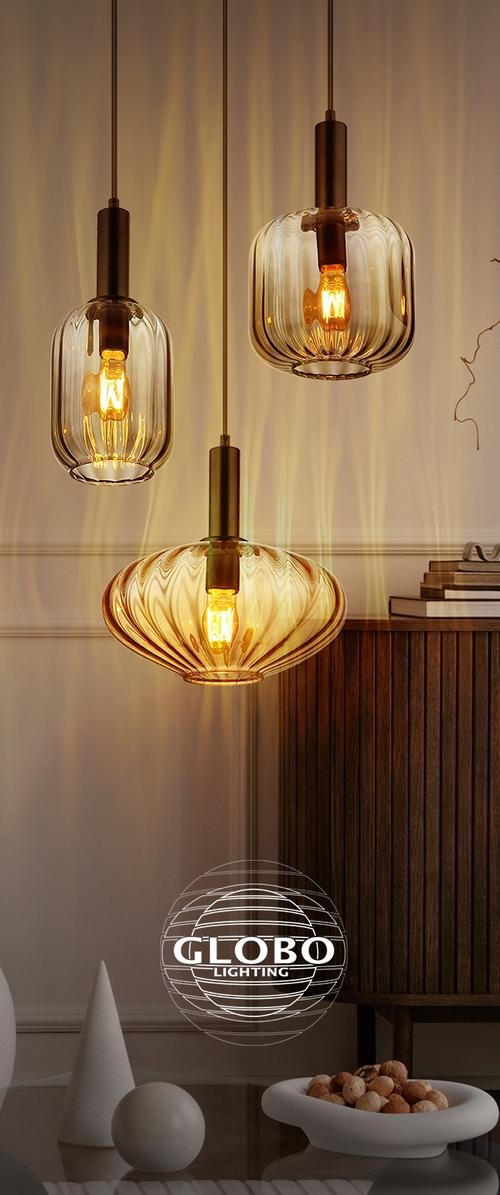 Tres lámparas colgantes de vidrio ámbar con bombillas expuestas iluminan un interior moderno con muebles de madera y objetos decorativos sobre una mesa.