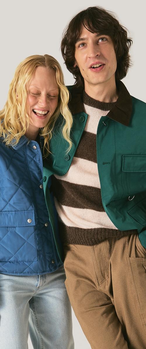 Femme souriante en veste matelassée bleue appuyée sur un homme portant une veste verte et un pull rayé, tous deux debout devant un fond uni.
