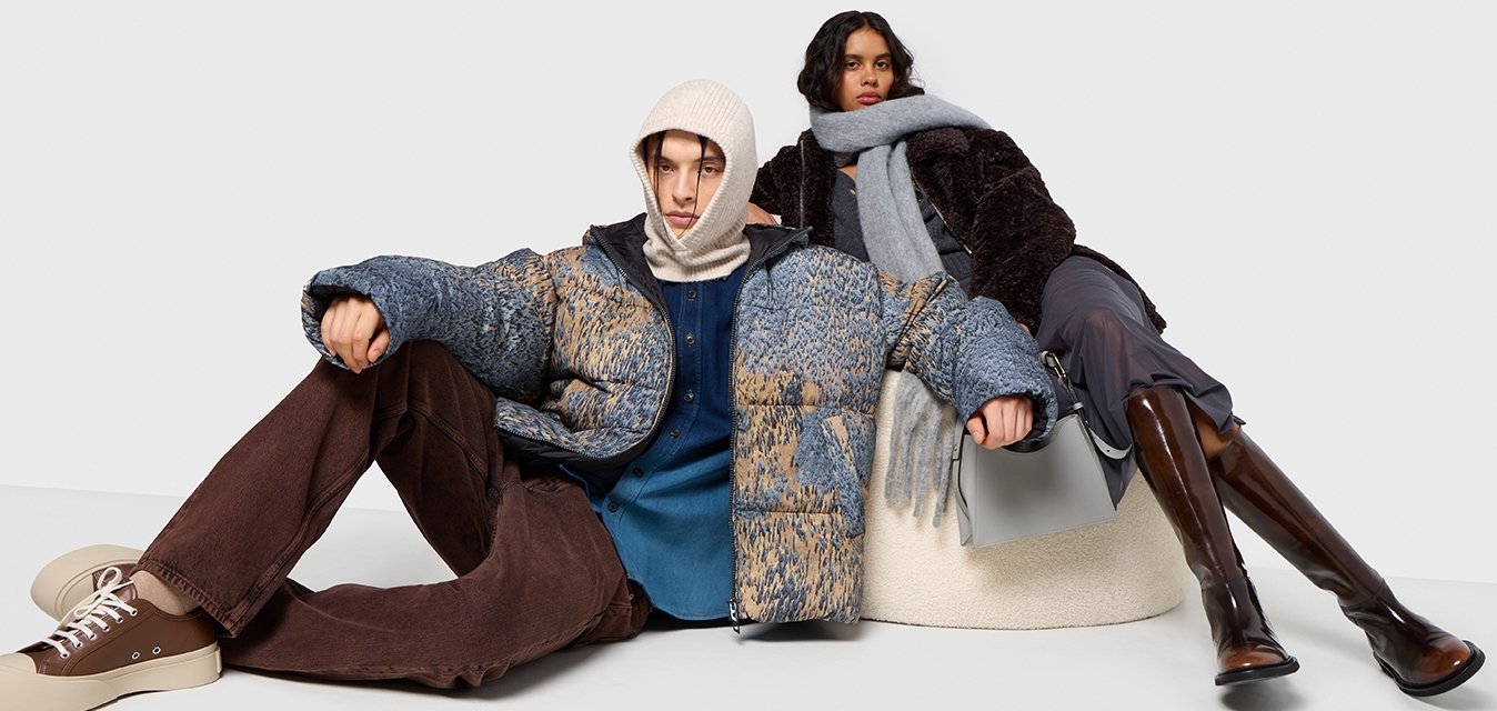 Pohodlná modro-hnědá bunda s podšívkou, šedý šátek, hnědé kalhoty s rozšířenými nohavicemi a hnědé boty. Měkké materiály a vrstvení oblečení v posedě.