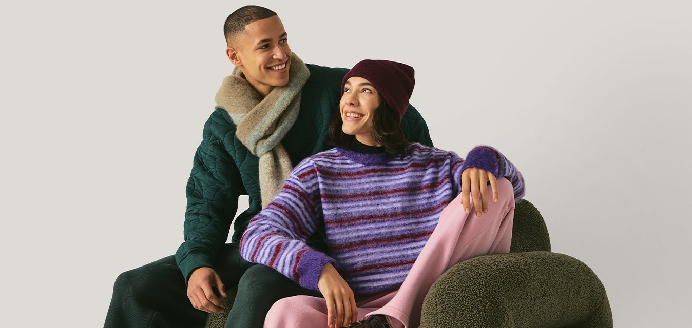 Usměvavý muž v zeleném svetru a šále sedí za ženou v pruhovaném fialovém svetru, růžových kalhotách a vínové čepici, která sedí na olivově zeleném křesle.