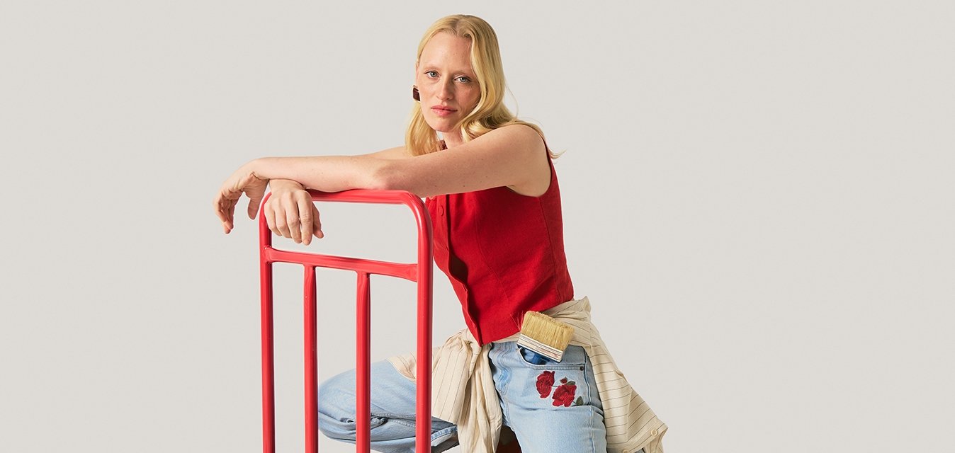 Žena v červeném topu a džínách klečí na černém plošinovém vozíku s červeným madlem na jednotném pozadí.