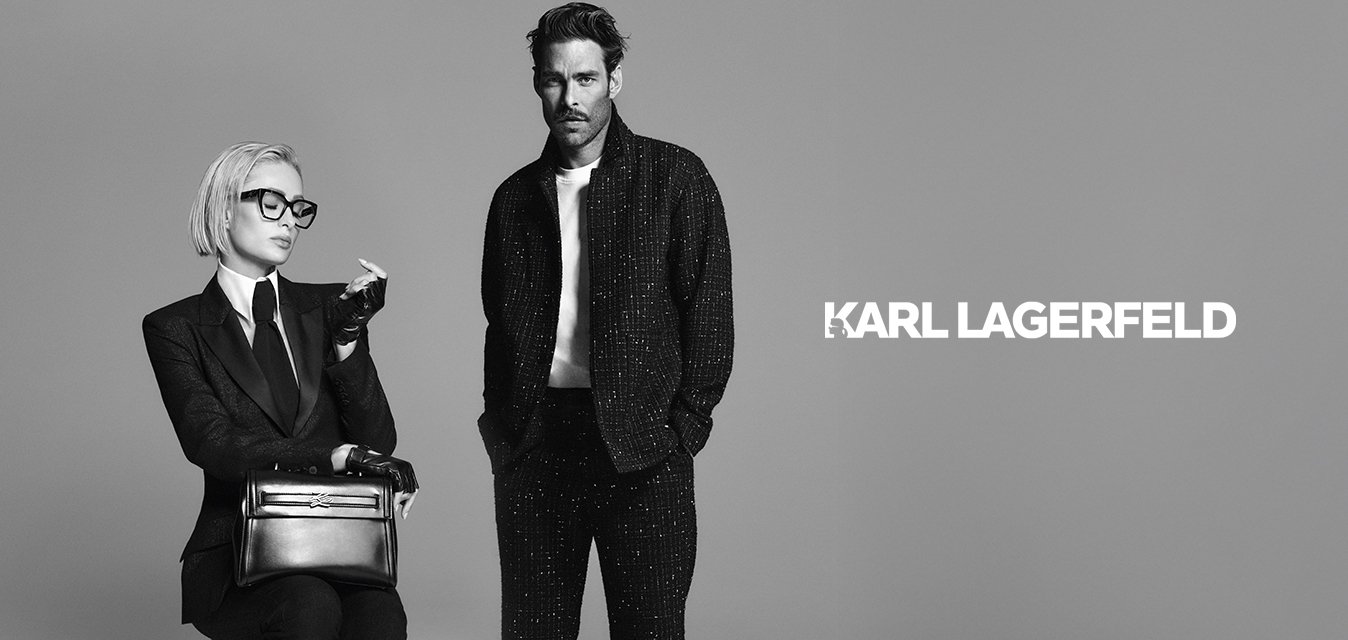 Žena s krátkými blond vlasy a brýlemi sedí na stoličce v obleku, rukavicích a mokasínách a drží kabelku; vedle ní stojí muž v neformálním tmavém oblečení.