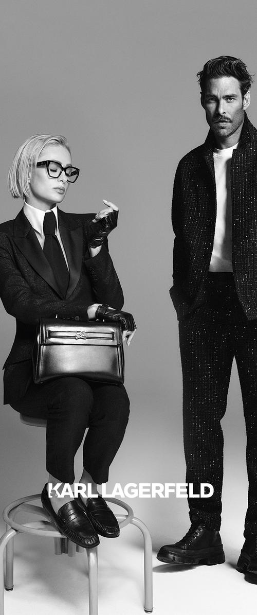 Image en noir et blanc mettant en scène une figure assise dans un costume sur mesure avec des gants et des lunettes, tenant un sac à main noir structuré, aux côtés d'une figure debout dans une tenue noire scintillante et des bottes.