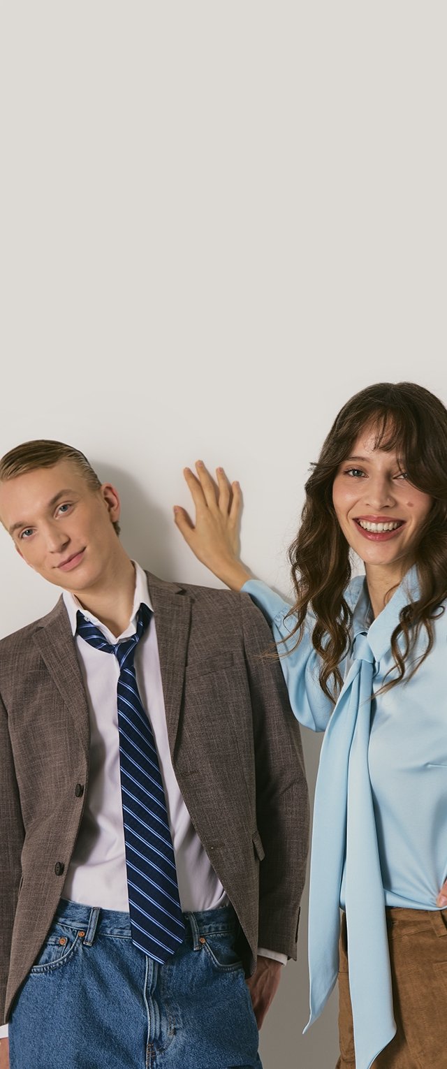 Deux personnes prennent la pose devant un fond neutre. L'une porte un blazer marron sur une chemise blanche et une cravate rayée ; l'autre porte un chemisier bleu clair avec un détail de cravate.