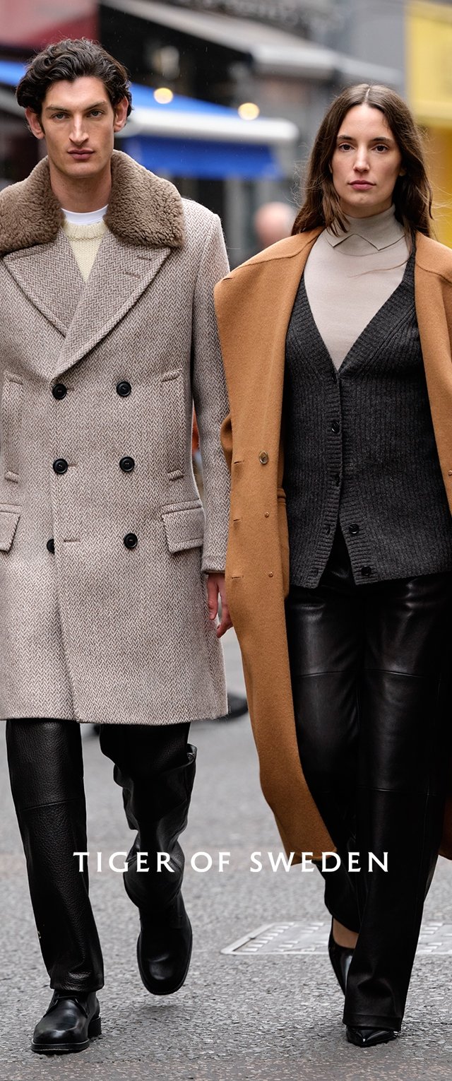 Dos modelos caminan en un entorno urbano. El hombre lleva un abrigo gris de espiga con solapas de piel y pantalones negros. La mujer lleva un abrigo marrón sobre un chaleco gris y pantalones de cuero.