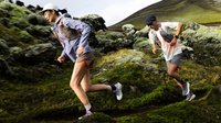 Zwei Trailrunner mit Kappen und Sportkleidung laufen bergauf auf einem moosbedeckten, felsigen Bergpfad.
