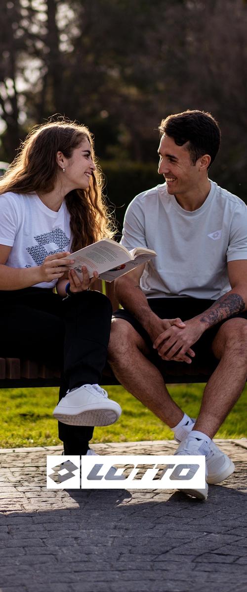 Dos personas están sentadas en un banco vestidas de forma casual. Una lleva una camiseta blanca con un estampado y pantalones negros, la otra lleva una camiseta gris claro y pantalones cortos. Ambas tienen calzado blanco.