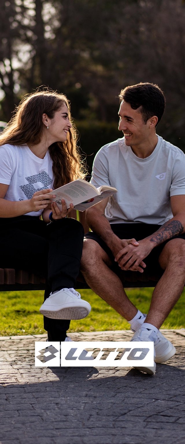 Dos personas están sentadas en un banco vestidas de forma casual. Una lleva una camiseta blanca con un estampado y pantalones negros, la otra lleva una camiseta gris claro y pantalones cortos. Ambas tienen calzado blanco.