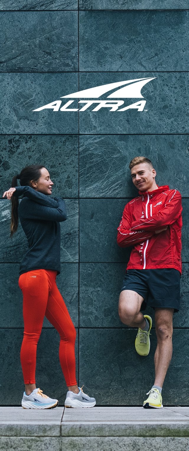 Twee personen staan tegen een donkere stenen muur. De ene draagt een zwarte trui en oranje leggings; de andere draagt een rode jas en shorts.