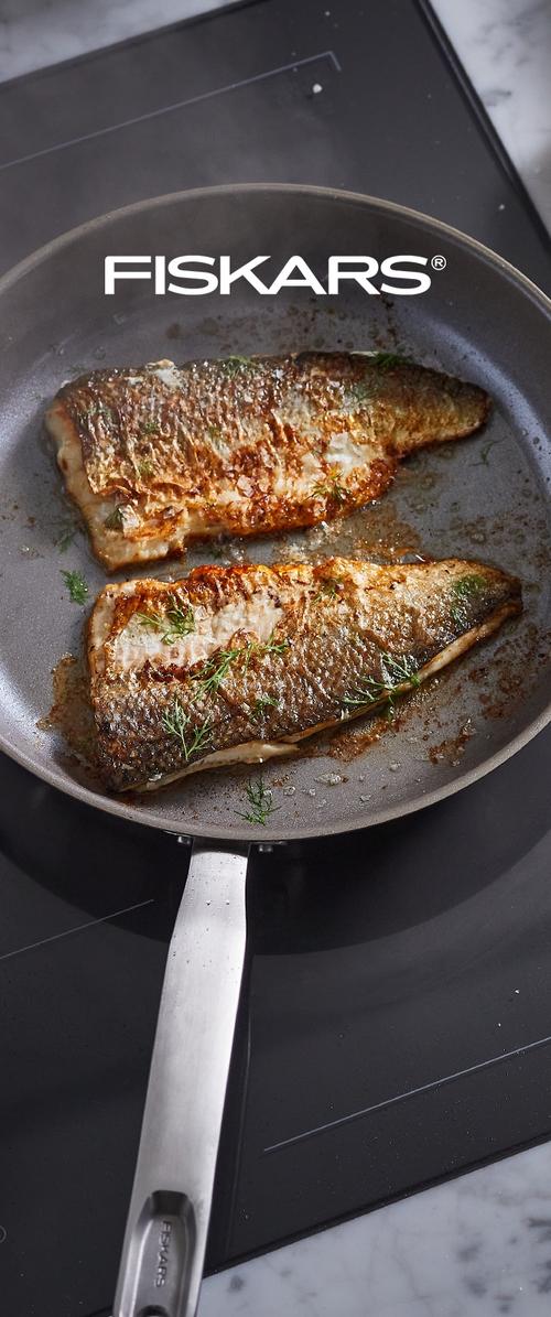 Dos filetes de pescado dorados y crujientes, adornados con hierbas, friéndose en una sartén antiadherente de Fiskars sobre una cocina.