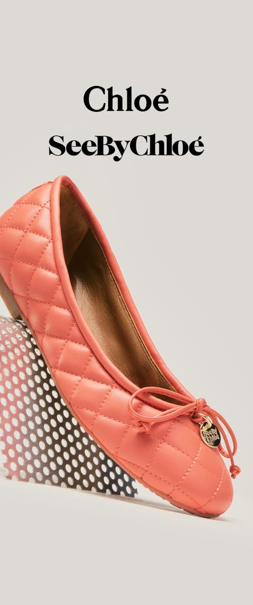 Pikowana koralowo-różowa balerina z kokardą i zawieszką SeeByChloé, ustawiona na perforowanym metalowym stojaku, z logo Chloé i SeeByChloé powyżej.