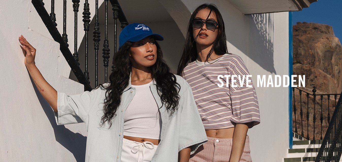 Dvě ženy v letních neformálních outfitech pózují u bílé budovy s modrým lemováním, venkovního schodiště a skalnatého pobřeží pod jasnou oblohou.