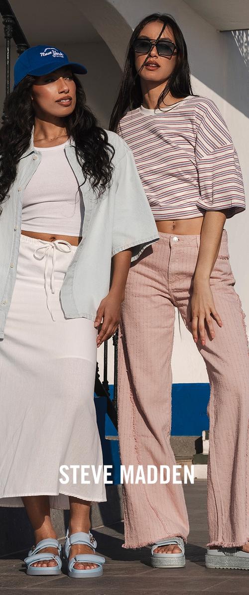 Zwei Frauen stehen im Freien und tragen lässige Sommeroutfits mit Sandalen; eine mit blauer Mütze und weißem Rock, die andere mit gestreiftem Oberteil und rosa Hose.
