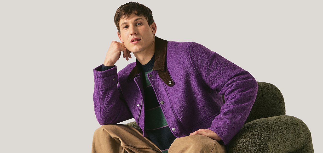 Mladý muž v fialové bundě a béžových kalhotách sedící na unikátně tvarovaném tmavě zeleném křesle na jednobarevném pozadí.
