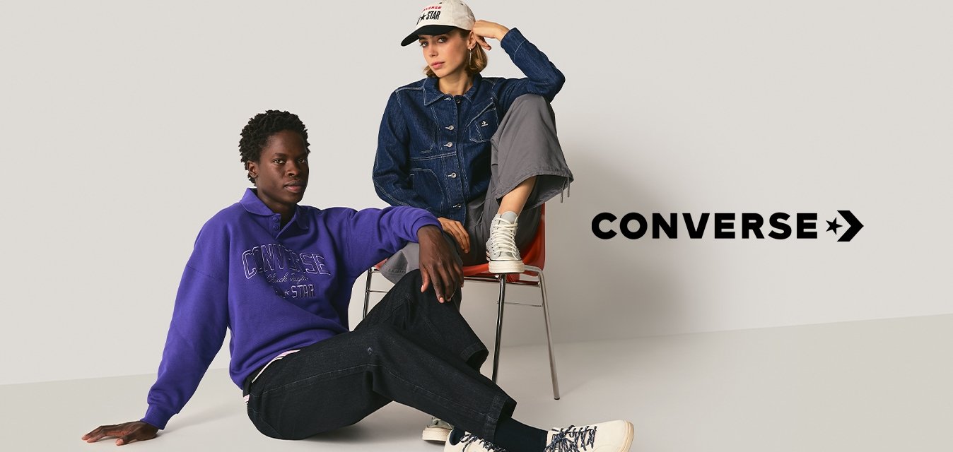 Dva lidé v běžném oblečení; jeden sedí na podlaze v fialovém svetru, černých kalhotách a bílých botách; druhý na židli v denimové bundě, šedých kalhotách a bílé čepici.